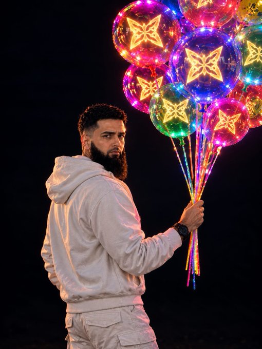 A man with a beard in a white hoodie stands against a dark background holding a bunch of colorful illuminated balloons featuring butterfly designs.