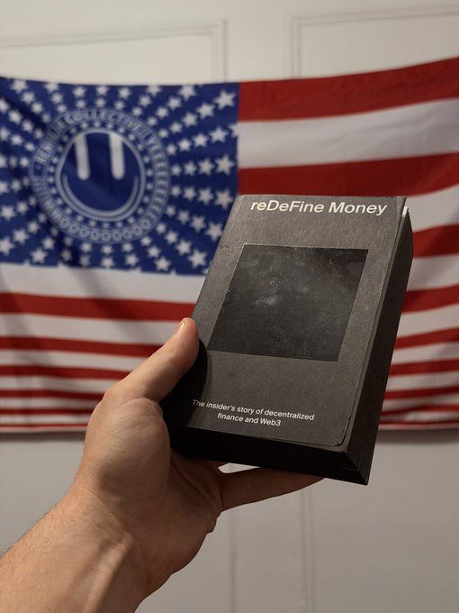 A hand holding a book titled 'reDefine Money: The insider's story of decentralized finance and Web3' in front of a US flag featuring a smiley face logo.