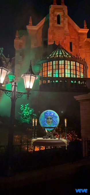 A nighttime scene featuring a crystal ball with a face inside, displayed on a table with candles in front of a gothic-style building illuminated with orange and green lights, branded with 'veve'.