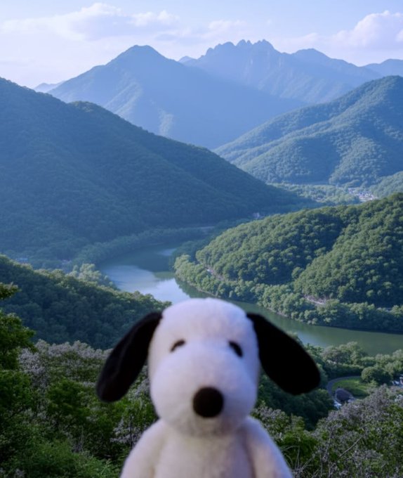 A plush toy of Snoopy in the foreground with a scenic view of green mountains and a winding river in the background.