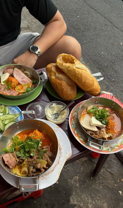 A person sits at a small outdoor table with two bowls of Vietnamese noodle soup (bún bò Huế) topped with beef and herbs, accompanied by a plate of crusty baguette bread and fresh cucumber slices.