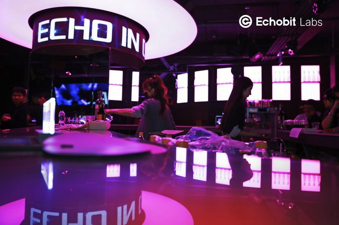 A dimly lit event space with purple and pink lighting, featuring an 'ECHO IN' sign and the 'Echobit Labs' logo, with people working behind a counter.
