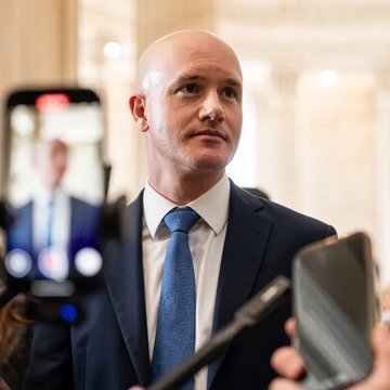 A bald man in a dark suit and blue tie is speaking to reporters, with a smartphone in the foreground recording him.