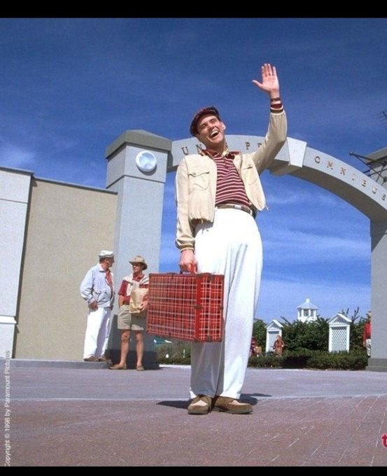 A man in vintage clothing waves joyfully while holding a red plaid suitcase at the entrance to Universal Studios, with the archway partially visible behind him.