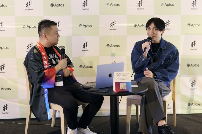 Two men sitting on chairs at a HeartPort World event in front of a backdrop covered with 'Aptos' logos, one holding a microphone while speaking to the other who also has a microphone.