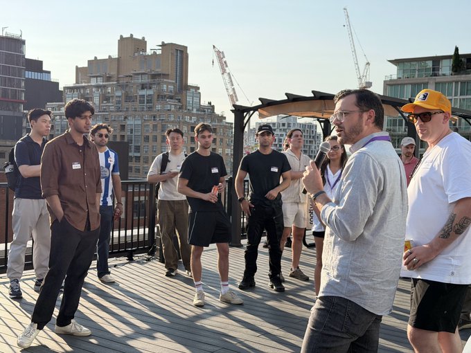 A man in a gray sweater speaks into a microphone on a rooftop terrace, addressing a group of casually dressed attendees at a networking event, with urban high-rises and construction cranes visible in the background. The setting reflects the post's description of a Rootly x OpenAI rooftop mixer for engineers, where $10k in Codex tokens were given away to participants discussing AI applications.