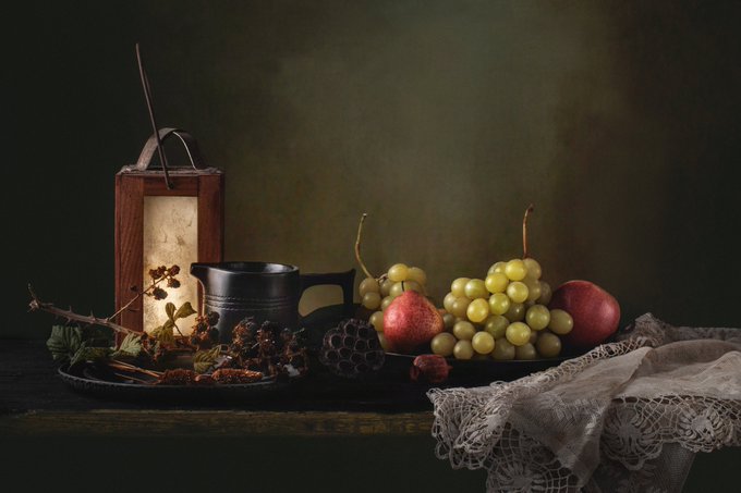 A moody still life painting-style image featuring a cluster of green grapes, apples, dried berries, and a vintage metal mug on a wooden surface with a white lace cloth.