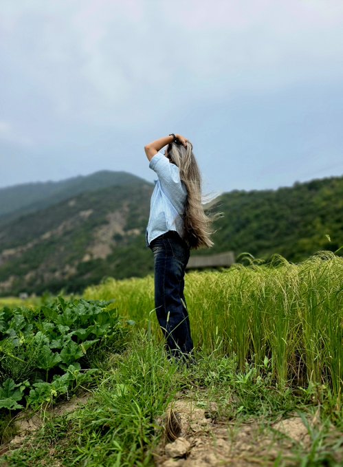 A person with long hair stands in a grassy field with their hands raised to their head, facing away from the camera toward distant mountains under a cloudy sky.
