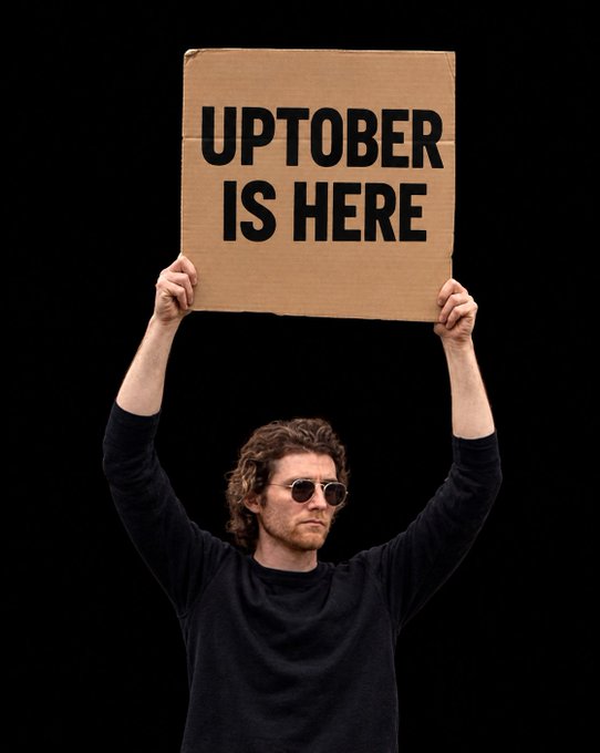A man in sunglasses holding up a cardboard sign that reads 'UPTOBER IS HERE', a play on the crypto trading term for October's typically bullish performance.