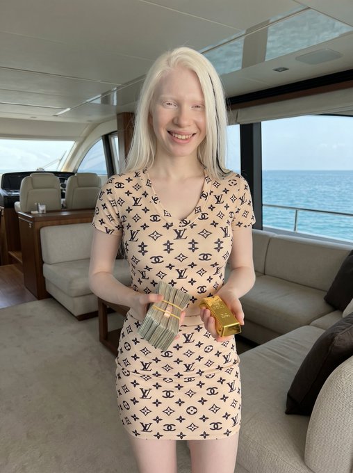 A smiling young woman with long blonde hair stands inside a yacht's luxurious cabin, holding stacks of cash and a gold bar while wearing a patterned dress, visually illustrating the cash and gold bar prize being promised in the post.