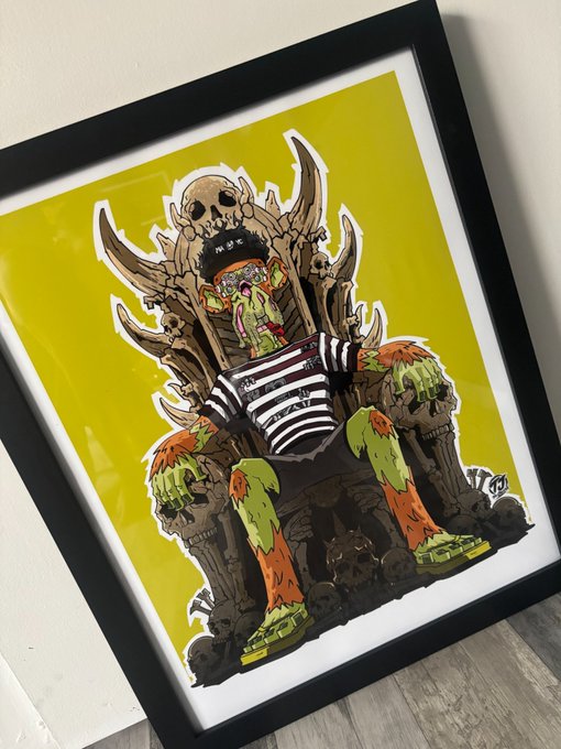 A framed piece of artwork featuring a cartoon character with a skull-topped throne, green slime, and bones set against a bright yellow background, leaning against a wall on a wooden floor.