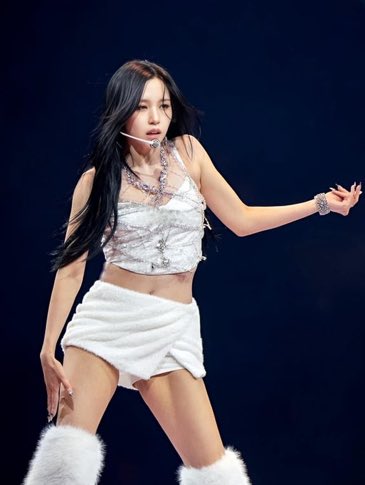 A female K-pop performer with long dark hair performs on stage wearing a white crop top with jeweled neckline, a white mini skirt, and fuzzy white boots.