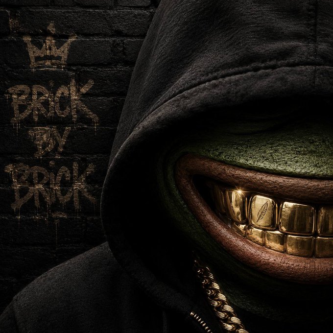 A close-up image of a hooded figure with green skin and a wide grin showing prominent gold teeth, with a gold chain visible, set against a dark brick wall tagged 'BRICK BY BRICK.'