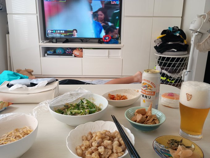 The image shows a table with various food items, including bowls of noodles, plates of food, and drinks such as beer. A person can be seen lying down on the floor next to the table, possibly enjoying their meal or taking a break from standing up. The scene appears to be set in a living room where there is a television turned on, displaying a sports event.