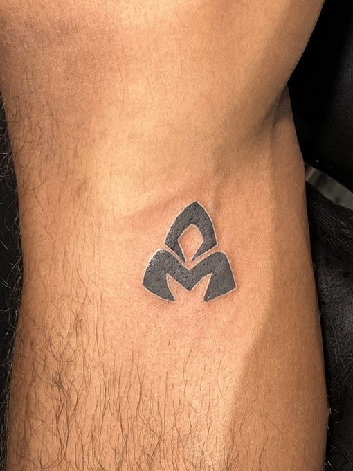 A small black tattoo of a stylized geometric symbol resembling a leaf or flame is inked on a person's ankle or wrist.