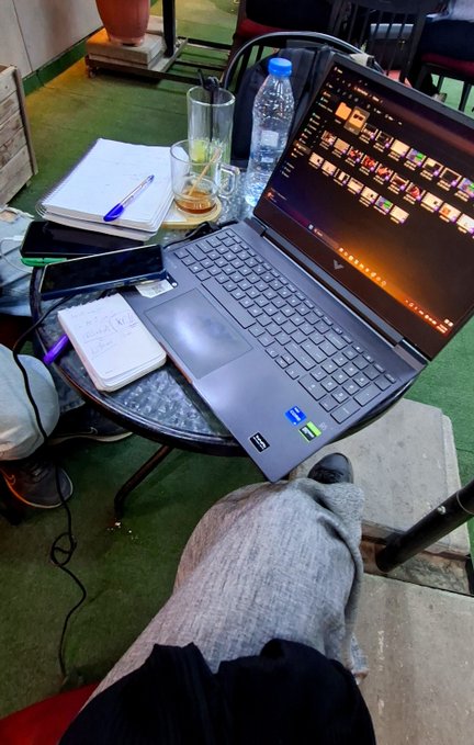 A first-person perspective shot of a laptop on a small round table at what appears to be a café, with notepads, a pen, drinks, and the person's legs visible.