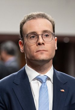 A man with short brown hair and wire-frame glasses wears a navy blue suit, white shirt, and light blue tie featuring a small American flag pin on the lapel. The image accompanies news of CFTC Chairman Mike Selig announcing the agency is ready to deliver its regulatory rules for the emerging frontier of finance.