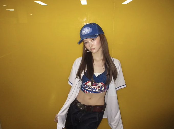 A young woman poses against a bright yellow wall wearing a blue baseball cap, a white baseball jersey over a cropped blue Dodgers-style top, and dark jeans with a decorative belt buckle.