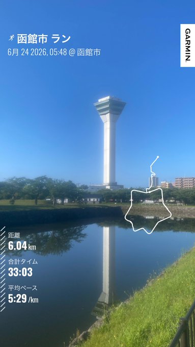 The image shows a tall white tower with a map of the area displayed at the top, along with a path or trail for hikers. The tower appears to be located near a body of water, possibly a lake or pond. The surrounding environment includes trees and grass, creating a picturesque setting for outdoor activities like hiking.