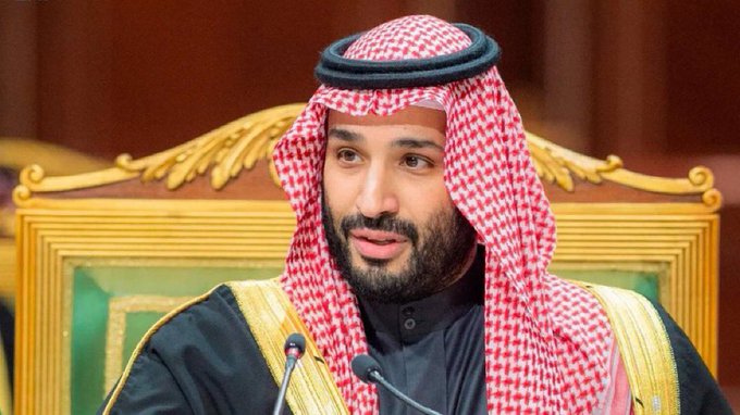 A bearded man wearing a traditional red and white checkered Saudi keffiyeh and black bisht sits in an ornate gold chair at a formal gathering.