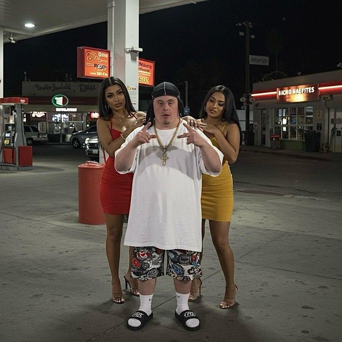 A man in a white t-shirt, bandana, gold cross necklace, and slides poses with two women in red and yellow outfits at a gas station at night.