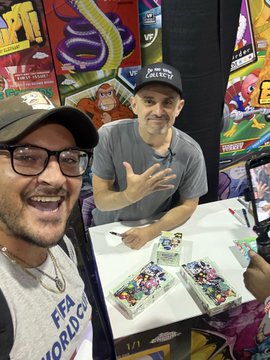 Two smiling men pose for a selfie at what appears to be a comic con signing event, with the man on the right seated behind a table displaying VeeFriends trading cards and artwork, while the "VEECON" logo is visible on the backdrop behind them.