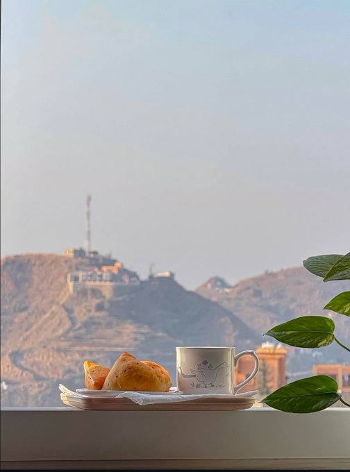 A peaceful scene shows a coffee cup and pastries on a saucer placed on a windowsill, with a green plant on the right and a mountain with a communications tower visible in the hazy background.