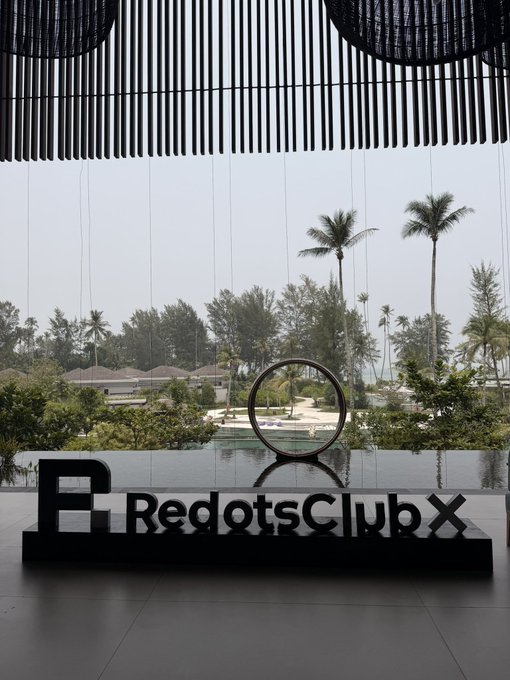 An interior shot of a building lobby featuring a black sign with white text reading 'RedotsClubX' next to a reflective pool with palm trees visible in the background.
