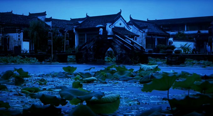 A twilight scene featuring traditional Chinese architecture with white walls and dark tiled roofs, an ornate stone arch bridge, and a pond filled with lotus leaves in the foreground.