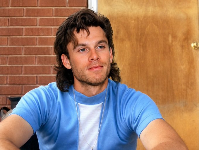 A young man with wavy brown hair wearing a light blue sweater over a white t-shirt, standing in front of a brick wall and wooden door.