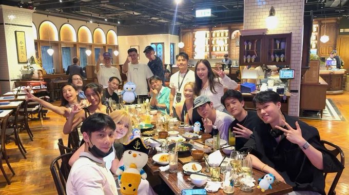 A group of about fifteen people pose cheerfully around a long dining table filled with plates, drinks, and Pudgy Penguins plushies in a warmly lit restaurant. Most are making peace signs and smiling for the camera, with one man in a white cap and another in a black mask seated in front. The post "Pudgy Penguins After Hours: Taipei Edition — Dinner with the Huddle" indicates this is a community meetup of Pudgy Penguins NFT fans in Taiwan, with the plush toys serving as a visual nod to the project's brand.