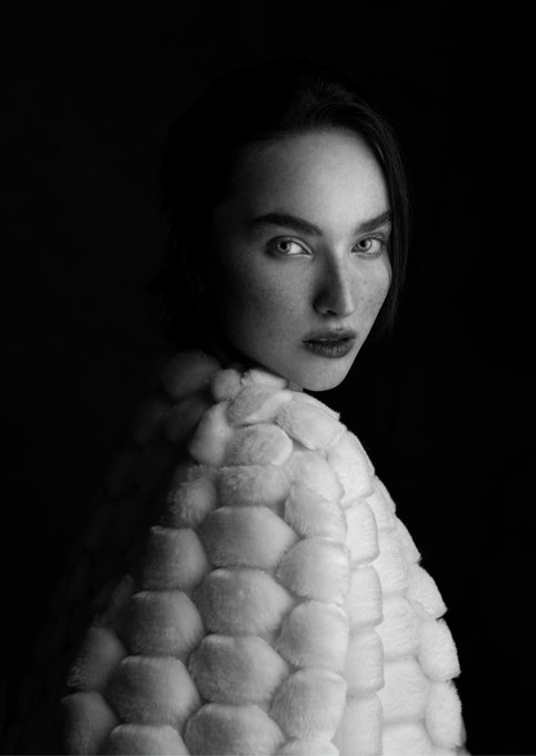 A black-and-white portrait of a woman with dark hair, freckles, and a serious expression, wearing a thick, textured white fur coat against a dark background.