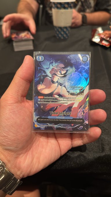 A hand holds a holo Pokémon TCG card featuring Primarina (Tidal Insight), with another stack of cards and a person in a blue patterned shirt visible in the background.