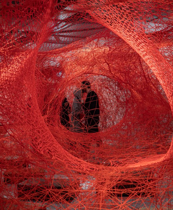 Two people embrace at the center of a large-scale art installation made of intricate, swirling red thread.