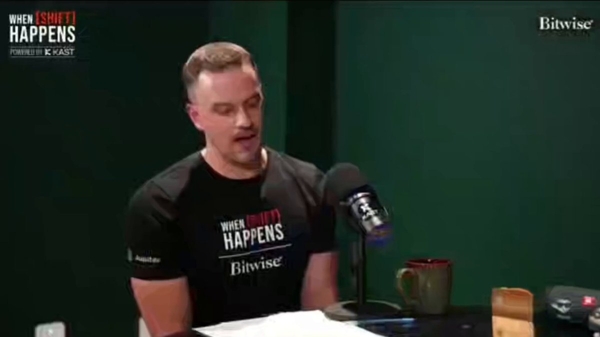 The image shows a man wearing a black t-shirt sitting at a desk with a microphone, appearing to be talking or giving a speech. The background of the image features a green wall, which contrasts with the man's dark clothing. There is also a cup placed on the desk in front of him. The post that accompanies this image discusses the concept of Hyperliquid and its implications for society. It highlights how massive perps demand can be brought into a regulated U.S. market by using technology to manipulate people's behavior, as seen in the man's speech or conversation with the microphone.