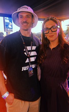Two people pose together indoors; the man wears a white bucket hat and black 'Just Do It' Nike shirt with a lanyard, while the woman wears glasses and a dark striped top.