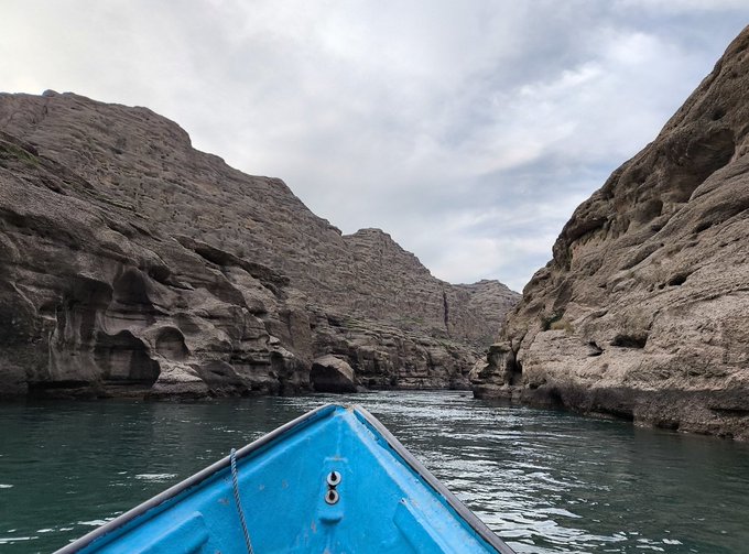 A bright blue boat navigates through narrow turquoise waters between towering, rugged limestone cliffs under a partly cloudy sky.