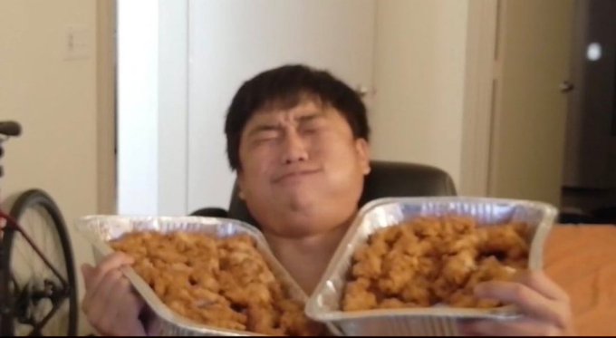 A man holds two large foil trays of fried chicken with a satisfied facial expression.
