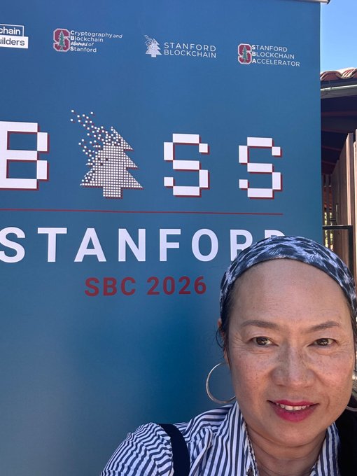 A woman wearing a blue-and-white striped headscarf and hoop earrings smiles in the foreground while posing at the Stanford SBC (Stanford Blockchain Accelerator) 2026 event, with a branded backdrop displaying "BASS STANFORD SBC 2026," "Cryptography and Blockchain at Stanford," and "Stanford Blockchain Accelerator" logos behind her—capturing her excitement as an attendee of the AI x Finance x Blockchain gathering.