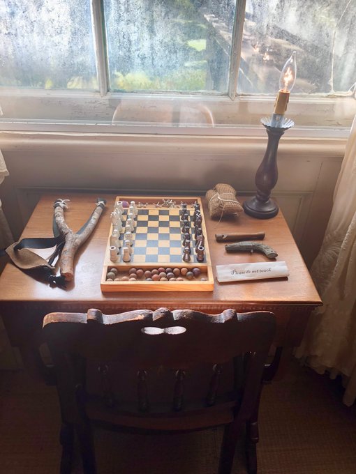 A small wooden table with a vintage chess set, sticks, twine, and a candle, with a small sign reading 'Please do not touch'.