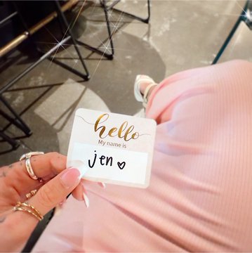 A hand with rings holds a name tag card reading 'hello My name is Jen ♡' with a pink dress visible in the background.