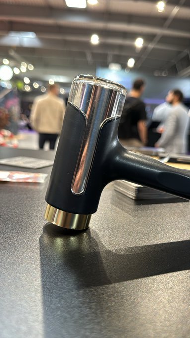 A close-up product photo of a black and chrome hair dryer sitting on a dark display table in what appears to be a trade show or retail environment with people visible in the blurred background.