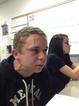 A teenage boy wearing a black 'MICHIGAN' hoodie sits in a classroom, looking toward the camera with a serious expression, while a girl sits at a desk beside him in front of a whiteboard.
