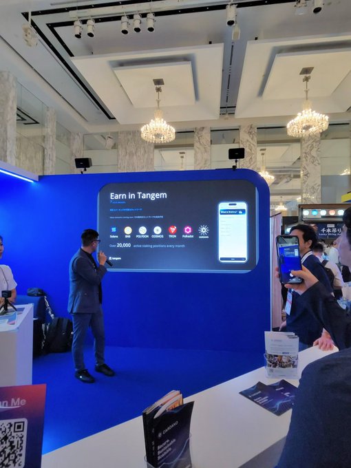 A man in a suit presents at a business event in an elegant venue, standing before a screen titled 'Earn in Tangem' that displays 'Over 20,000 active staking partners every month' alongside various cryptocurrency logos.