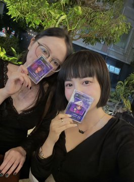 Two women in black tops posing together at night, each holding up a small card with purple and white design near their faces.