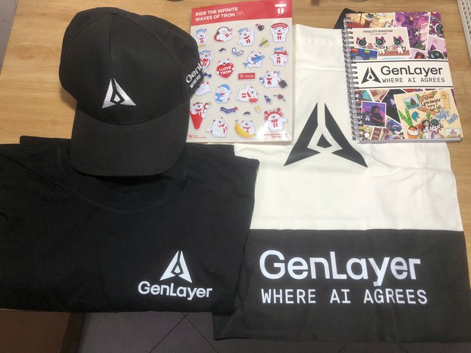 An assortment of GenLayer merchandise laid out on a wooden surface, including a black cap, black and white t-shirts printed with 'GenLayer WHERE AI AGREES', a sticker sheet, and a spiral notebook featuring anime-style artwork.