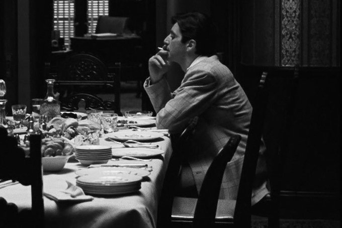 A black-and-white photograph of a man in a light suit smoking a cigarette while sitting at a table set with dishes and glasses.