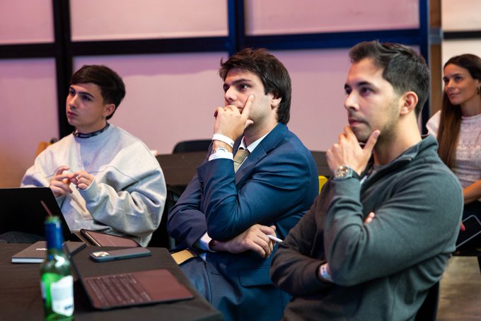 Three men seated in what appears to be a conference or classroom setting, with one in a blue suit thinking with his hand on his chin, alongside a younger man in a gray sweatshirt.