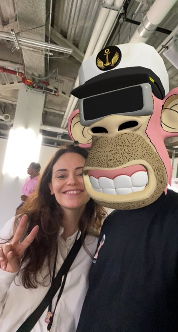 A woman smiling and making a peace sign while posing next to a person in a pink monkey mascot costume wearing VR goggles and a captain's hat.