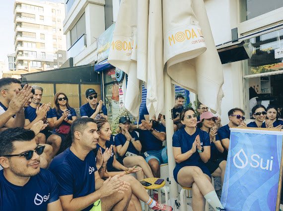 A group of people in matching blue 'Sui' t-shirts sit outdoors applauding, with umbrellas displaying 'MODA' and 'CAN DEMIR' behind them.